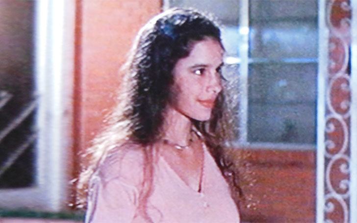Christin Hinojosa is smiling, standing in front of the orange house, has brown wavy hair, wearing a necklace, pink v neck shirt.
