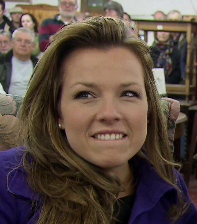 Christina Trevanion smiling while biting her lower lip with some people in the background and wearing a shirt under a purple coat, and pearl earrings