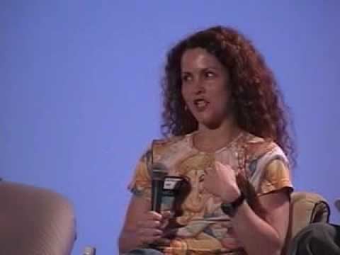 Christine McGlade is talking during an interview while sitting on a couch and holding a microphone, with curly hair, wearing a watch and a multi-colored shirt.
