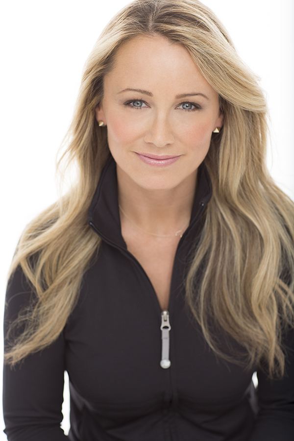 Christine Taylor with a tight-lipped smile and blonde wavy hair while wearing earrings and a black jacket