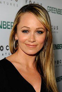 Christine Taylor with a tight-lipped smile and blonde straight hair while wearing hoop earrings and a black blouse with a low-cut neckline