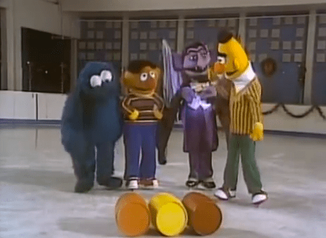 In a large room with blue walls with white tiles at the back, from left, Cookie monster is serious, standing looking down hands down, has large white eyes and dark blue body, 2nd from left, Emie is serious, standing with his hand on his waist, has black hair brown head wearing a stripe long sleeve a blue pants and white shoes, 3rd from left, Count von Count is serious, standing with his hand on his chest, has black hair a long eyebrows and fangs, wearing white top with black cape and a gray pants with black shoes, at the right, Bert is serious standing, looking at his right, has black hair, yellow body orange nose, wearing a yellow striped long sleeve with white turtleneck and a green pants with white shoes, in front is an Orange, yellow and brown barrel at the floor,