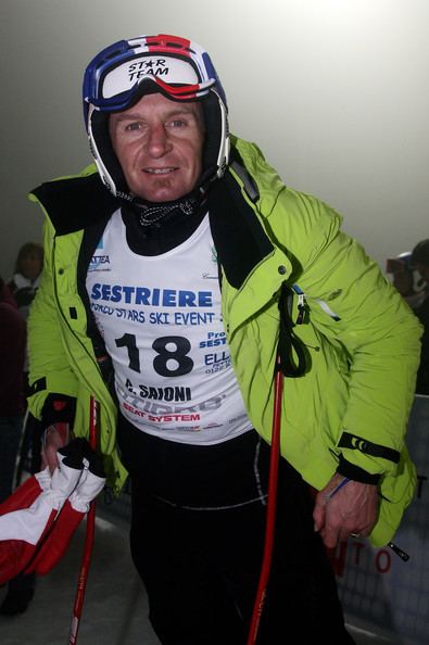 Christophe Saioni smiling with ski poles and gloves in his hand. Cristophe wearing a ski suit with words written like "SESTRIERE" and the number 18 under a yellow-green jacket, a colored helmet, black pants, and a finger ring