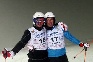 Christophe Saioni and Bruno Kernen embracing each other and holding ski poles during the 5th World Stars Ski Event in Sestriere in Turin, Italy. Christophe wearing a black and white ski suit with the printed word "SESTRIERE" and the number 18 and goggles while Bruno also wearing a blue and white ski suit with the printed word "SESTRIERE" and number 17 and both are wearing gloves and a helmet