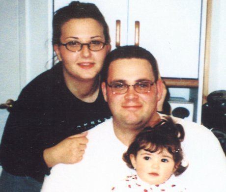 Christopher, Jaime, and Sophia Amoroso are smiling. Christopher is wearing eyeglasses and a white shirt, Jaime wearing eyeglasses and black long sleeve shirt while their daughter Sophia is with short curly hair and wearing a red and white shirt.