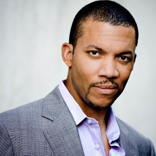 Christopher B. Duncan smirks while wearing a purple shirt and a gray coat in his pictorial