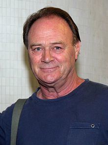 Christopher Cazenove wearing a blue shirt with a small pocket and a sling on his right shoulder.