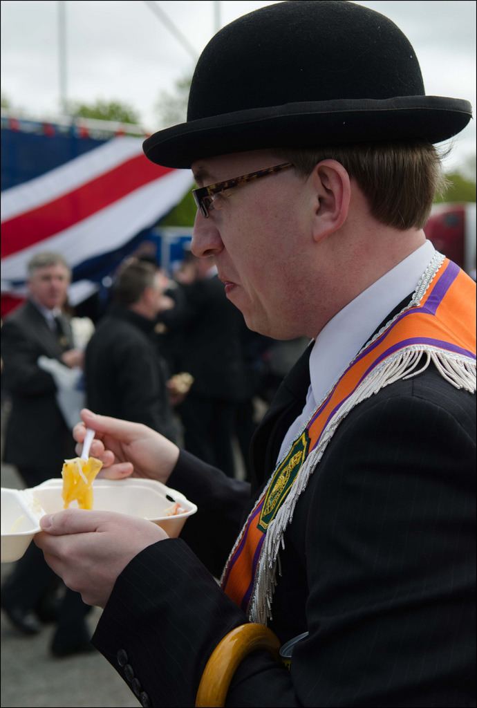 Christopher Stalford was looking from afar, has a black hat, black eyeglasses, holding a fork of food in a styrofoam food pack  with people in the background, and wearing a white long sleeve with a black necktie under a black suit with an orange collarette