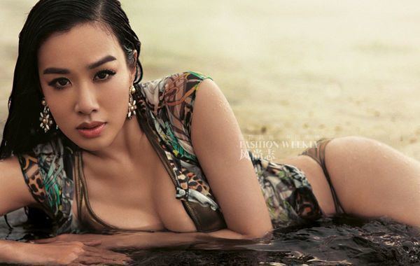 Christy Chung lying on the sand while wearing a brown and blue blouse, brown bikini, and earrings