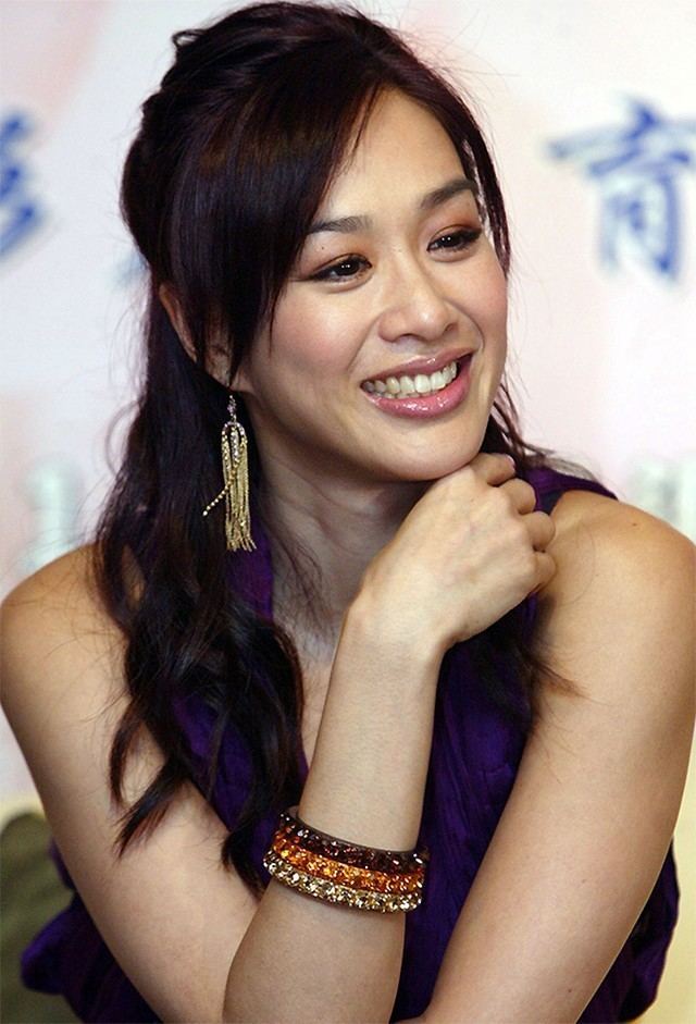 Christy Chung smiling while hand on her chin and wearing a blue sleeveless blouse, earrings, and bracelets