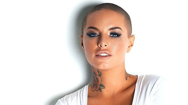 Christy Mack with a fierce look, bald head, and tattoos on her neck while wearing a white t-shirt