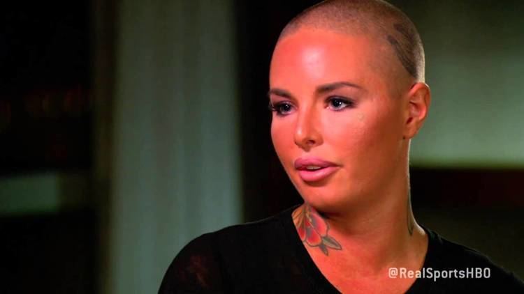 Christy Mack talking to the interviewer with a tattoo on her neck, a serious face, and a bald head while wearing a black blouse