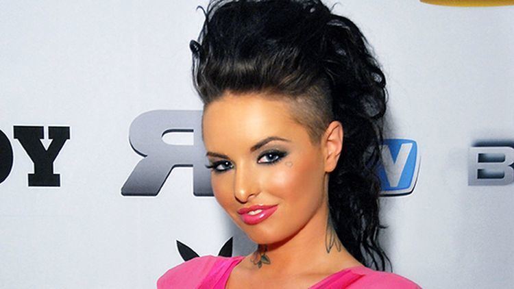 Christy Mack smiling while at the AVN Expo, Las Vegas, Nevada, with tattoos on her neck and an undercut hairstyle, and she is wearing a pink body fit sleeveless blouse