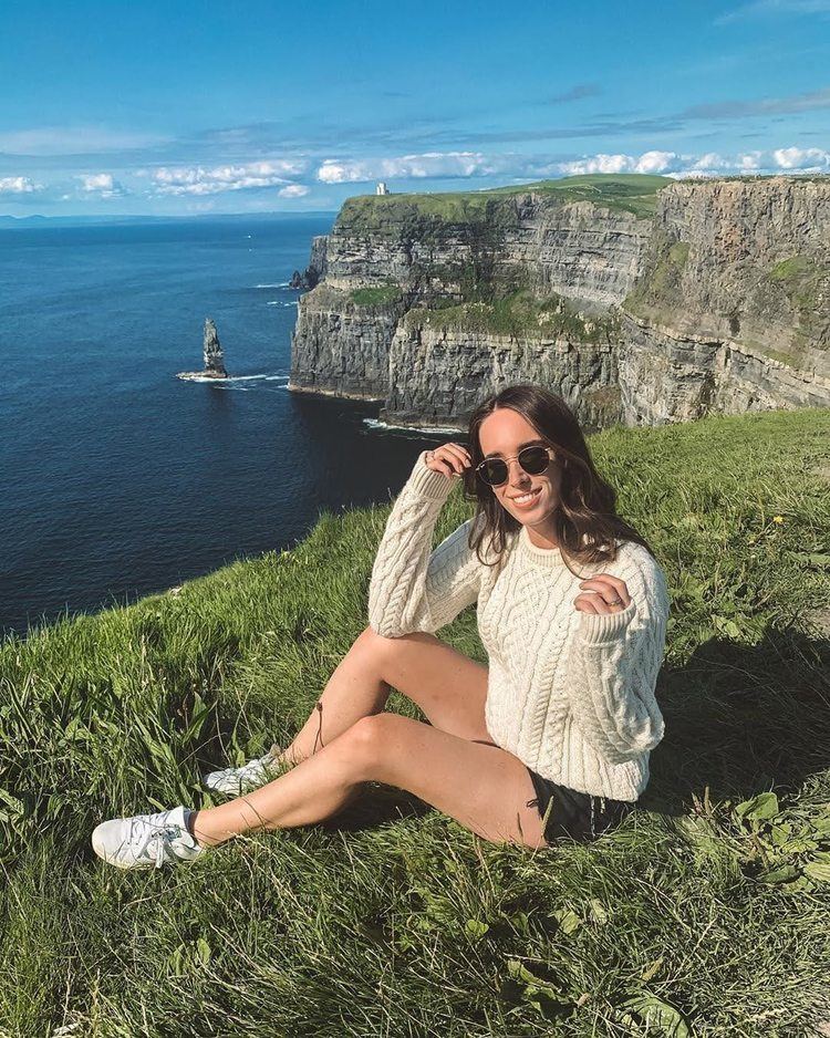 Ciara O'doherty smiling while sitting on the grass with a sea and mountain view and she is wearing sunglasses, white sweater, and shorts