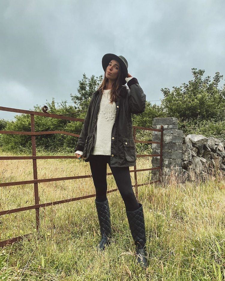 Ciara O'doherty holding a black hat while standing on grassland and wearing a black jacket, white blouse, black pants, and black boots