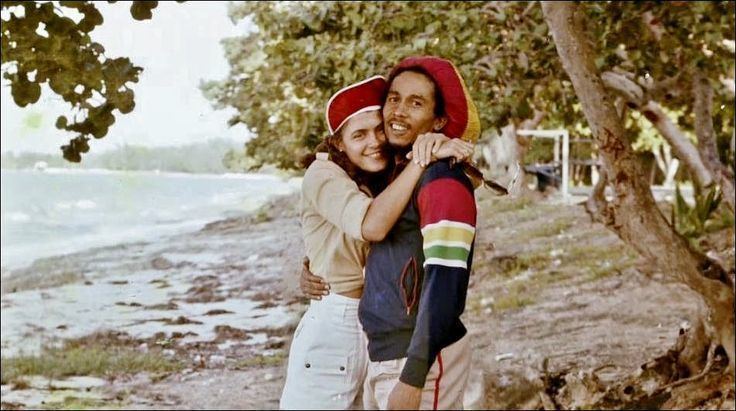 Cindy Breakspeare wearing a red cap, a brown top, and white shorts while hugging Bob Marley wearing a yellow and red hat and a colorful jacket.