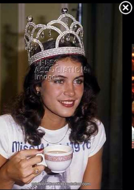 Cindy Breakspeare with a smiling face and curly hair, wearing a crown, a white shirt, and holding a cup of coffee.