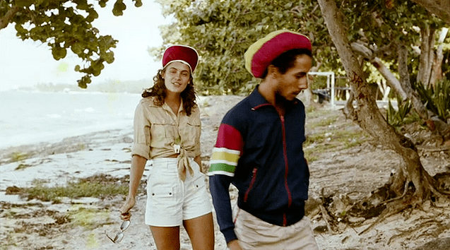 Cindy Breakspeare with curly hair, wearing a red cap, brown top, and white shorts together with Bob Marley wearing a yellow and red hat and a colorful jacket.