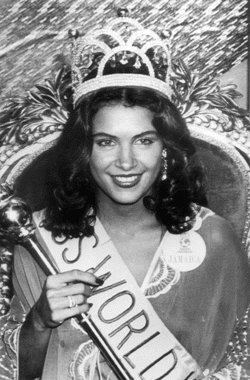 Cindy Breakspeare with a smiling face and curly hair, wearing a crown and a sash while holding a scepter.