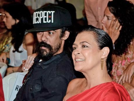 Cindy Breakspeare with smiling face, wearing earrings and a red dress with Bob Marley with a mustache and beard, wearing a cap and a black shirt.