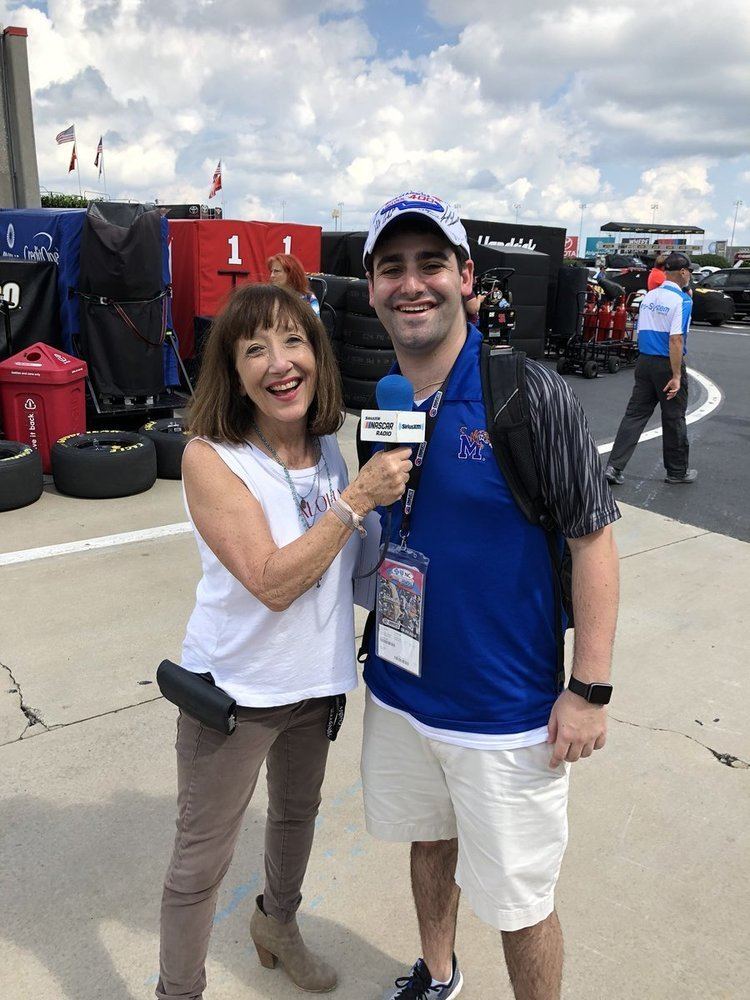 Claire B Lang happily interviewing Daniel Barrach during a racing show and wearing a white sleeveless shirt and brown pants.