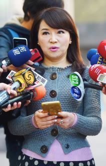 At the back is a man standing, has black hair, wearing a blue jacket and black pants. Clara Chou is serious, speaking, half mouth open, holding her phone with both of her hands, standing with microphones around her, has long brown hair wearing a purple shirt under a blue-green crochet long sleeve, white pearl earrings, a silver ring, and a black polka dot skirt.