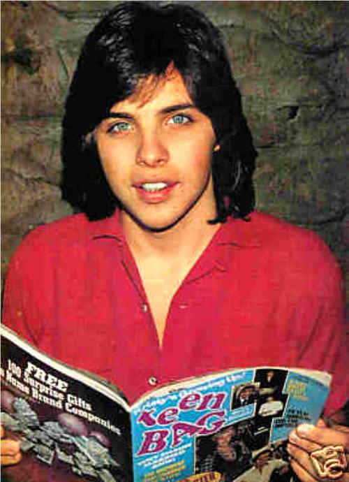 Young Clark Brandon smiling while holding a magazine, with long hair, and wearing a red polo shirt.