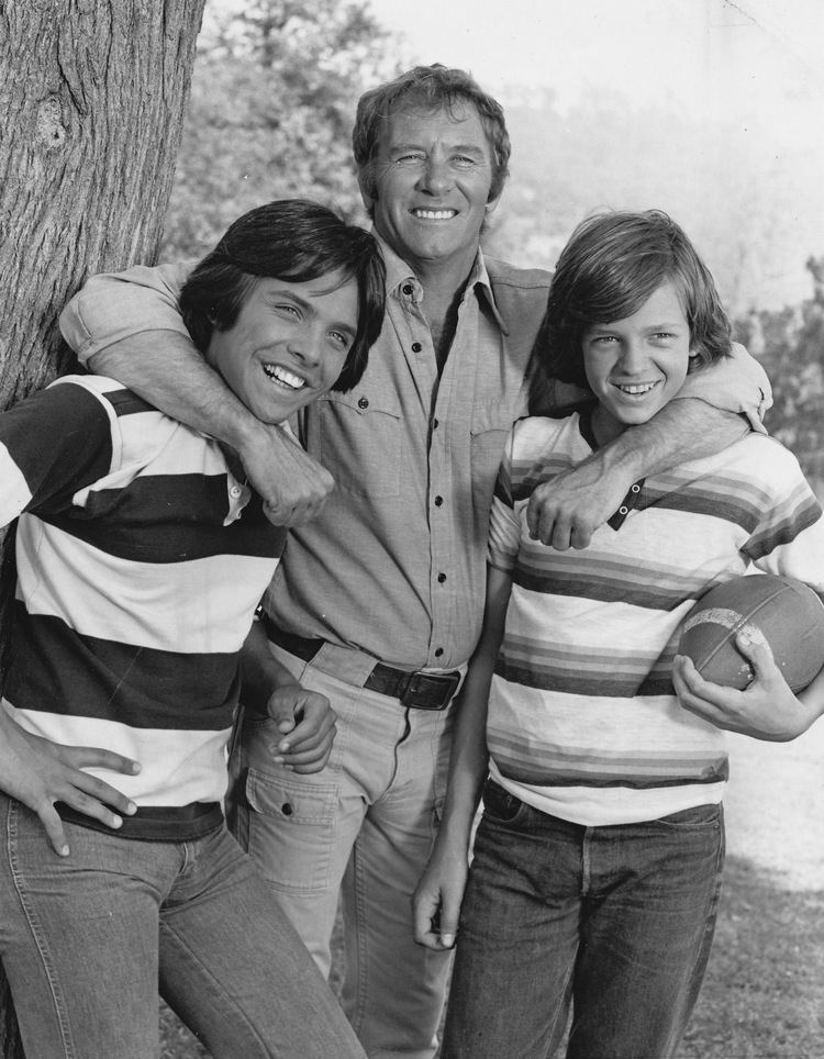 Clark Brandon, Peter Brandon, and Jimmy McNichol are smiling. Clark wearing a black and white polo shirt, Jimmy holding a football and wearing a striped polo shirt, and Peter wearing gray long sleeves.