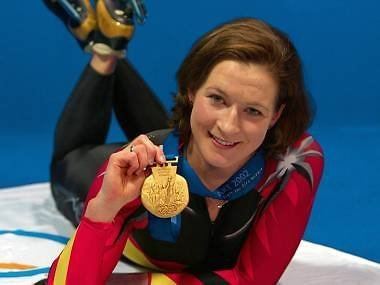Claudia Pechstein smiling while lying on the floor and holding a gold medal, with short hair and wearing a multi-colored bodysuit and skating shoes.