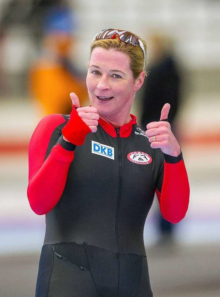 Claudia Pechstein is smiling while doing a thumbs-up hand gesture, with blonde hair, wearing sunglasses on her head, and a black and red bodysuit with logos.