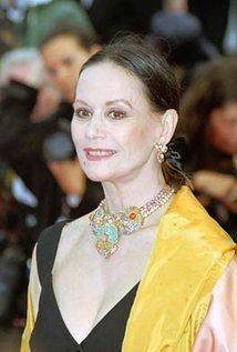 Claudine Auger smiling while wearing a black blouse, yellow blazer, necklace, and earrings