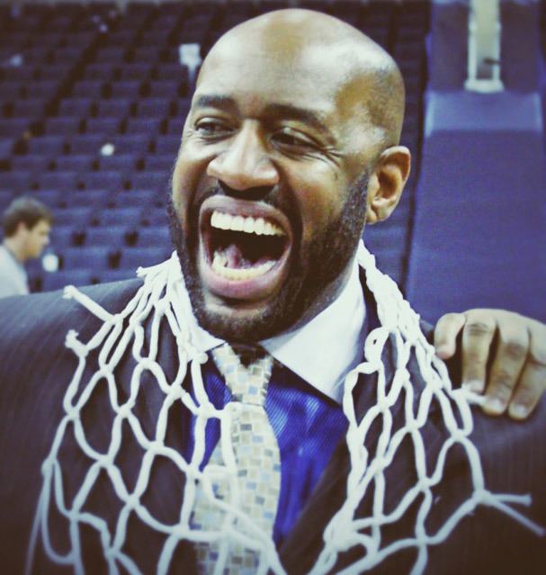 Cleo Hill laughing while wearing a black coat, blue long sleeves, necktie, and a basketball net