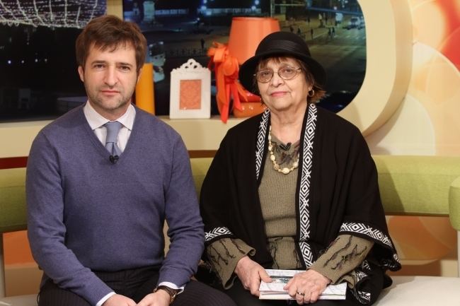 Călin Vieru and Raisa Vieru with serious faces while sitting on a couch. Călin is wearing a purple sweatshirt over white long sleeves and a purple tie while his mother is wearing a black hat, eyeglasses, and black and white blazer over a green long sleeve blouse.