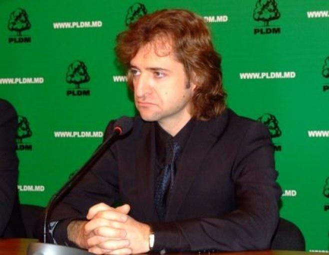 Călin Vieru with a sad face in front of a microphone while his hands are on the table, wearing a black coat over black long sleeves and black tie.