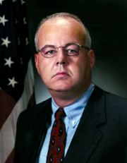 In black backdrop with the USA flag at the back, Cofer Black is serious, has white hair wearing eyeglasses, sky-blue polo shirt with red necktie under a black coat.