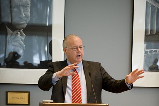 In a conference room, with gray wall and framed photos on the wall and a table with a mic on top, in front, Cofer Black is serious, speaking, hands up open, standing, has white hair wearing eyeglasses, sky-blue long sleeve polo with red striped necktie under a black coat.