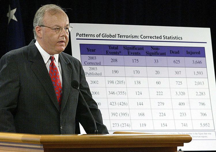 In black background with a USA flag at the back, Cofer Black is serious, speaking into the mic, standing in a conference room with a large presentation at his left side of the Patterns of Global Terrorism: Corrected Statistics, has white hair, gray eyes, wearing eye glasses & white polo with red necktie under a black coat.