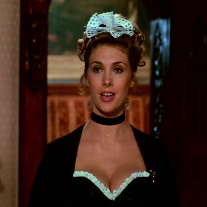 Colleen Camp as Yvette, smiling, with curly hair, in a scene from Clue, a 1985 American black comedy-mystery film. She is wearing a light blue headdress, a black choker, and a black and white dress with a visible cleavage.