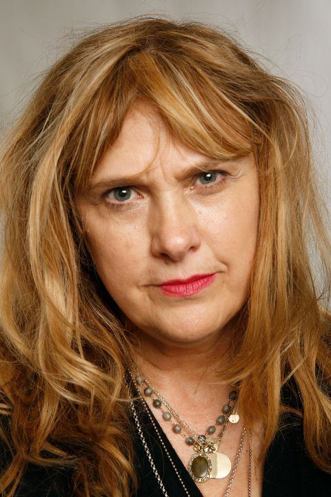 Colleen Camp with a serious face, blonde hair, wearing necklaces and a black shirt.