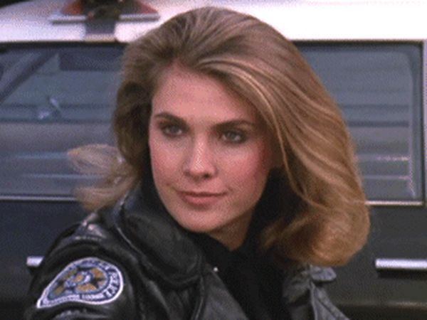 Colleen Camp smiling with a serious face, and blonde hair, in a movie scene from Police Academy 2: Their First Assignment, a 1985 American comedy film. She is wearing a black police uniform.