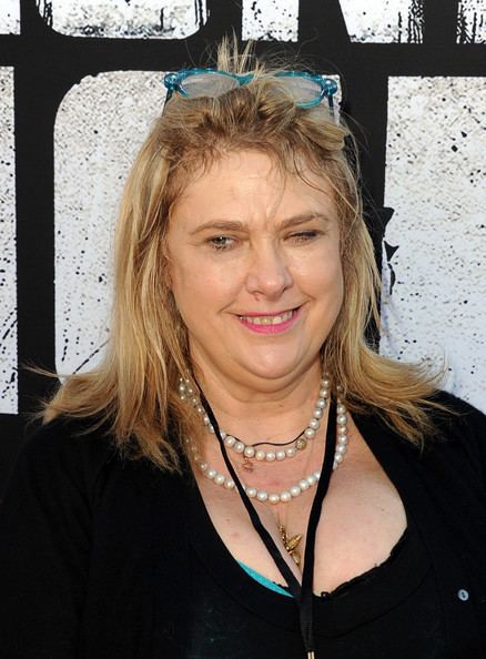 Colleen Camp smiling, with blonde hair, wearing eyeglasses on her head, necklaces, an ID, and a black blazer over a black shirt with a visible cleavage.