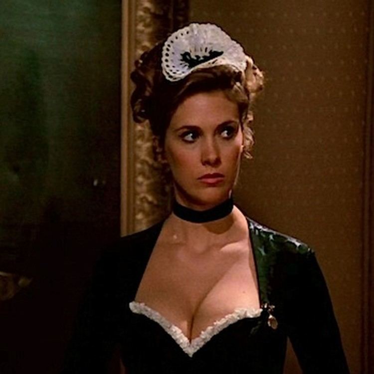 Colleen Camp as Yvette, with a serious face while looking at something, with curly hair, in a scene from Clue, a 1985 American black comedy-mystery film. She is wearing a white headdress, a black choker, and a black and white dress with a visible cleavage.