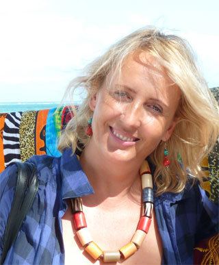 Corinne Hofmann smiling, with blonde hair and wearing a colorful necklace and a checkered blue polo shirt.