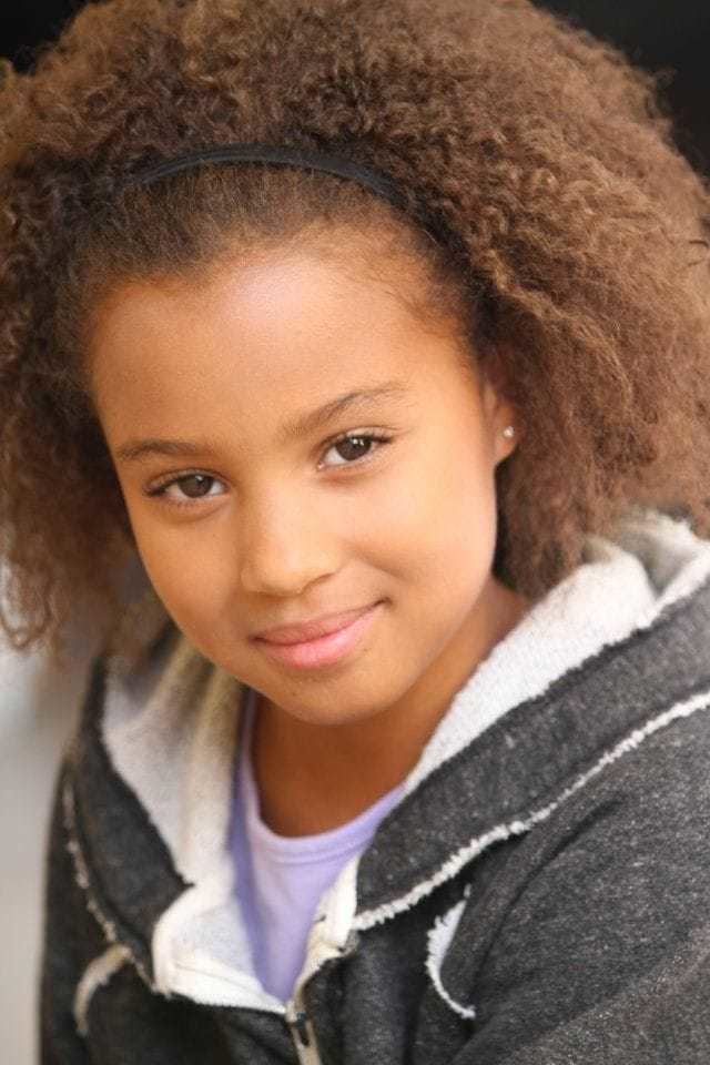 Corinne Massiah smiling with curly hair while wearing a headband and a purple blouse under a gray and white jacket