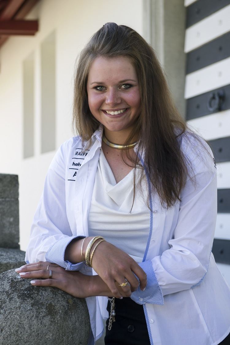 Corinne Suter smiling with her hair down, wearing a gold necklace, bracelets on her right wrist, and rings on her both fingers, wearing a white top under a white long sleeve