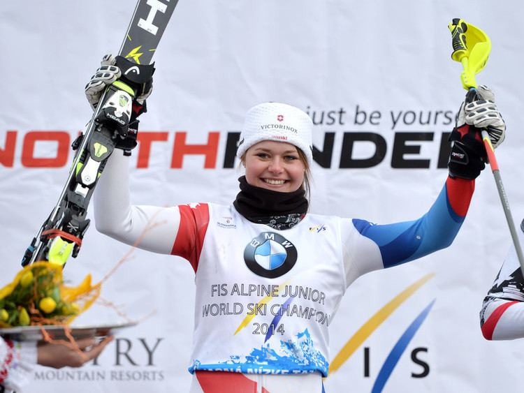 Corinne Suter smiling while raising her ski racer gears, wearing a white bonnet, a black scarf, gloves, and a white with red and blue long sleeve with a “FIS ALPINE JUNIOR WORLD SKI CHAMPION 2014” print on it