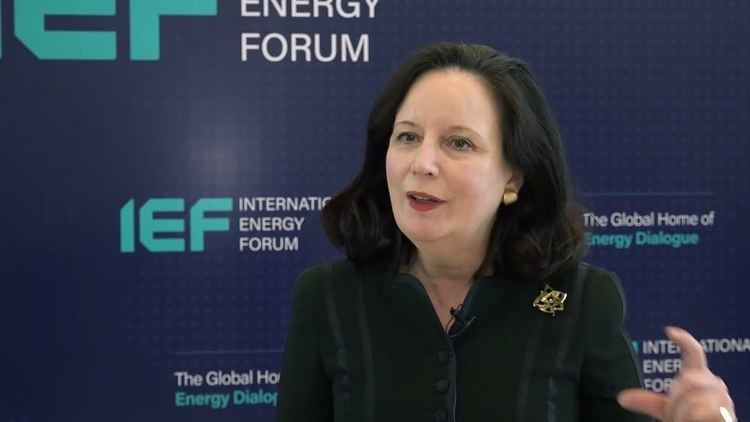 In the event of the International Energy Forum, at the back is a dark blue “IEF” printed backdrop, in front, Cornelia Meyer is serious, speaking, mouth half open, has black hair wearing a gold earring, a black blouse with a black mic and gold brooch on the left chest.