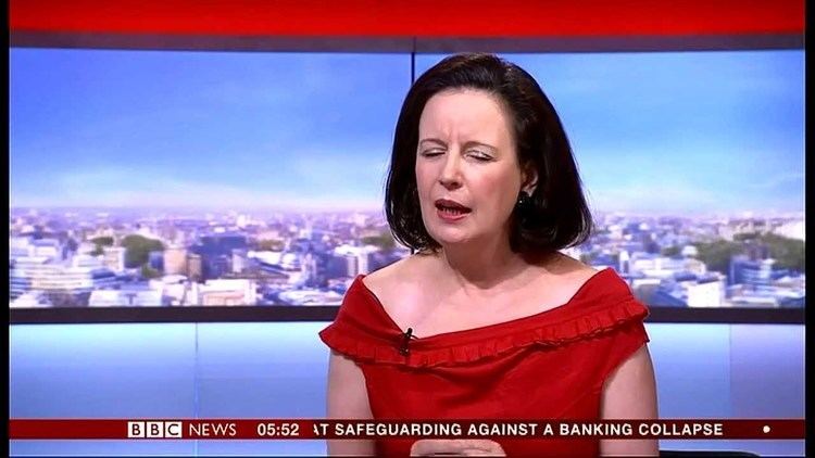 In the BBC news with a flat screen at the back broadcasting a city building, in front, Cornelia Meyer is serious, speaking, mouth half open, has black hair wearing earrings, a red dress with a microphone on the right chest, at the bottom is the BBC NEWS lines with time “05:52 at SAFEGUARDING AGAINST A BANKING COLLAPSE”