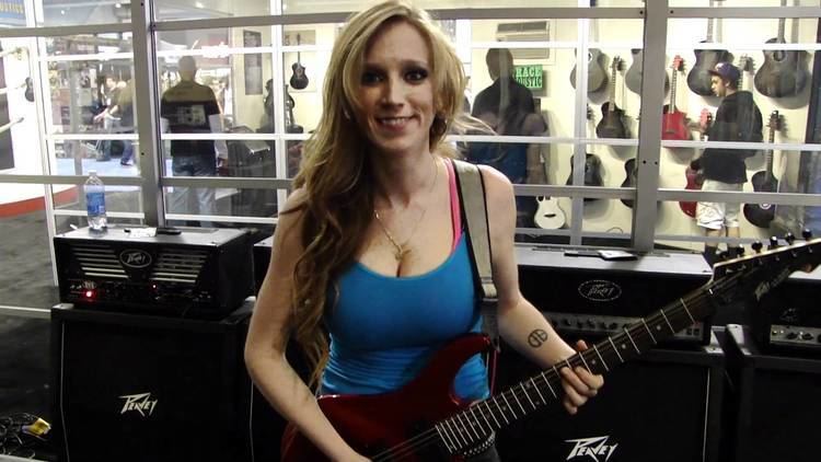 Courtney Cox smiling while holding an electric guitar and wearing a blue sleeveless top