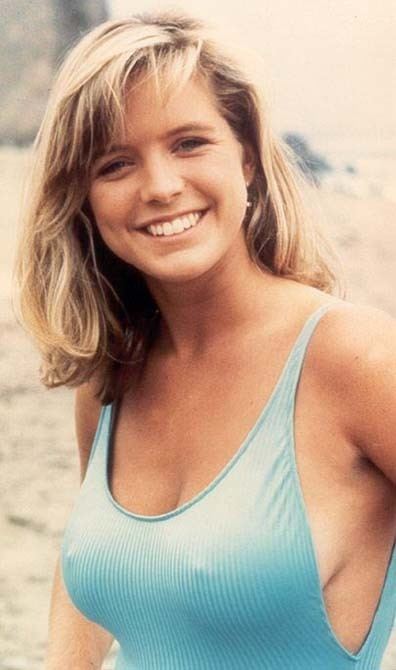Courtney Thorne-Smith smiling, an American actress with medium-length wavy blonde hair, has earrings, braless, wearing a light blue low-round neck one-piece with visible cleavage.